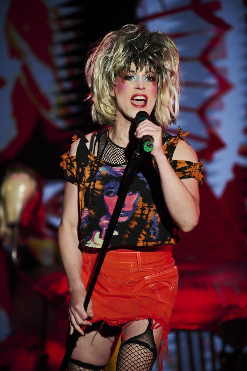 Performer in blonde wig, heavy makeup, black mesh top, orange skirt, and fishnets sings into a microphone on a stage with red lighting.