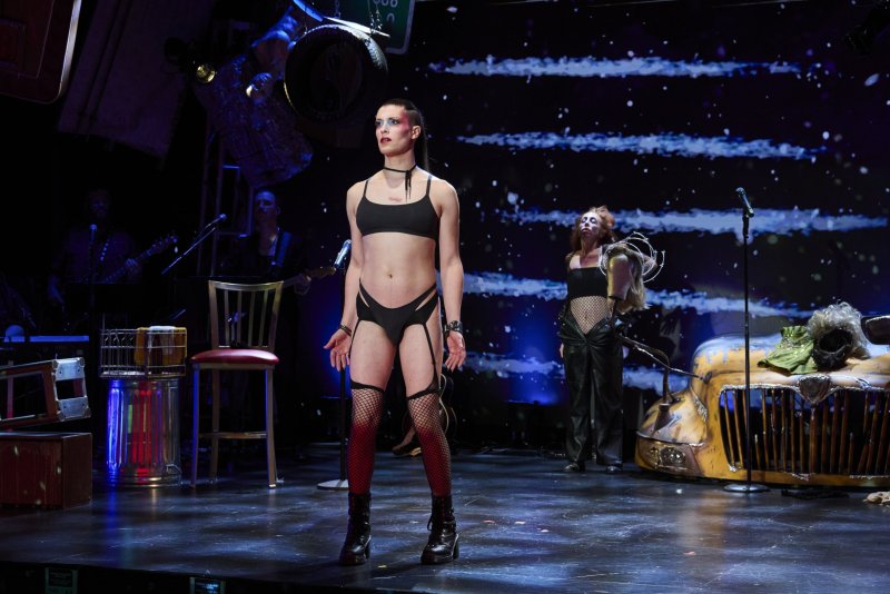 Performer in black bra, panties, fishnets, and boots on a dark stage with a starry background, a band, and a yellow car prop.