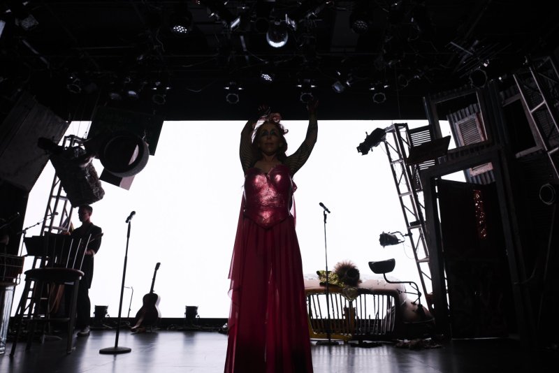 Drag queen in a shimmering pink dress with arms raised, performing on a dark stage with bright backlighting and musical instruments.