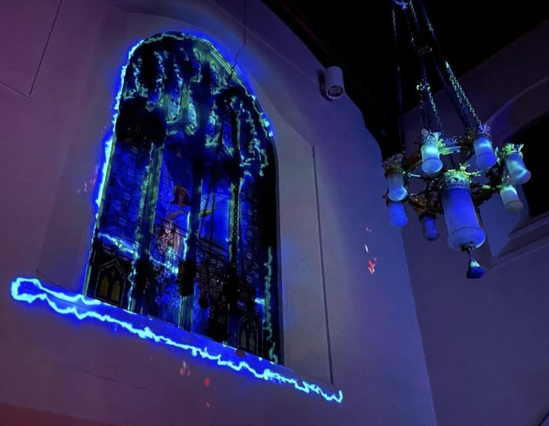 Stained glass window glowing with blue light, an angelic figure at its center, next to a chandelier with blue-lit bulbs.