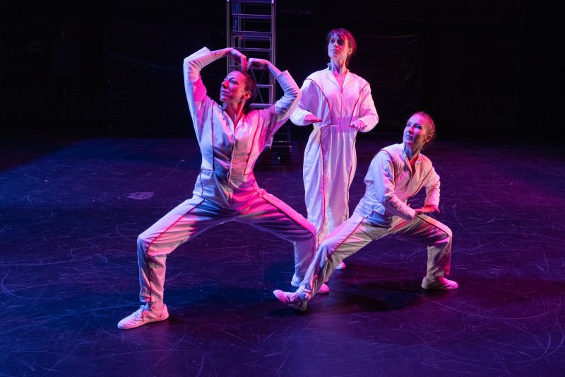 Three performers in white jumpsuits on a dark stage with pink and blue lighting. Two crouch, one stands.