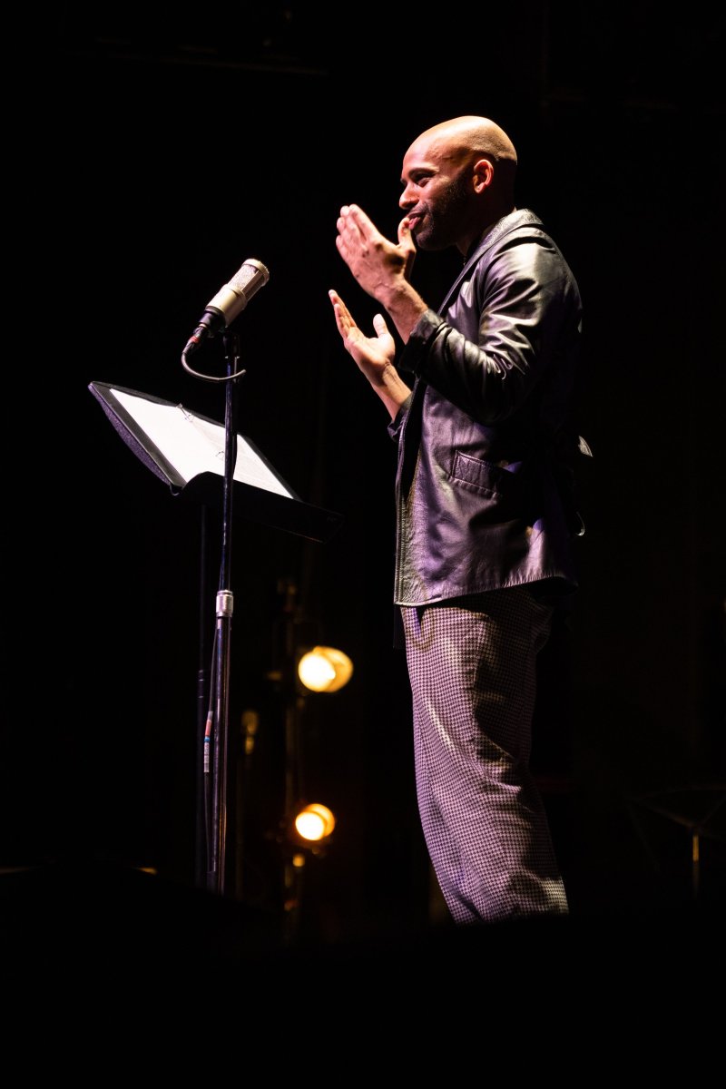 Bald man in leather jacket and plaid pants speaks into a microphone, hands gesturing, on a dark stage with warm spotlights.