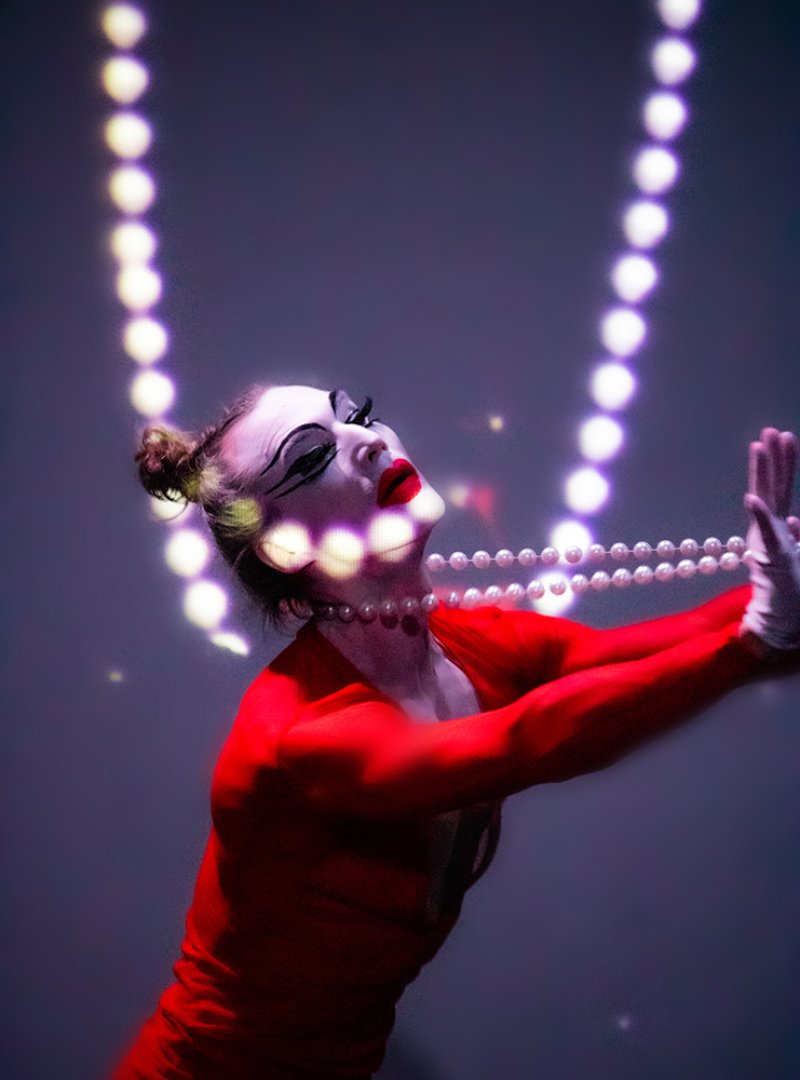 Performer in red, white gloves, and dramatic makeup, holding a pearl necklace, illuminated by vertical strings of light.