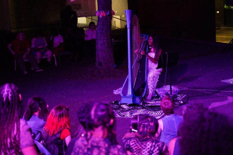 Outdoor performance bathed in purple light. A performer in a feathered dress plays a blue harp to a seated audience. A tree with decorations stands nearby.