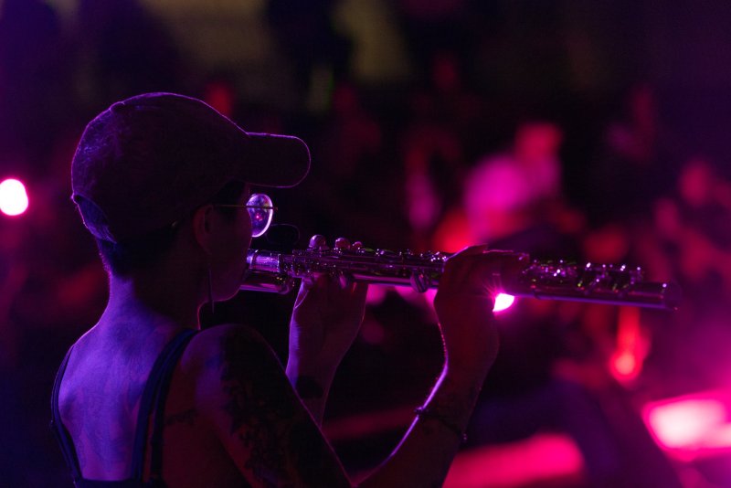 Back view of a musician in a cap and glasses playing a flute, lit with purple and pink stage lights, tattoos visible on their arm and back, with a blurred crowd in the background.