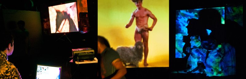 Multi-screen video installation. A man in a patterned shirt sits at a laptop. Screens show a shirtless man with a sheep on a yellow background, and silhouetted figures bathed in blue and green light.