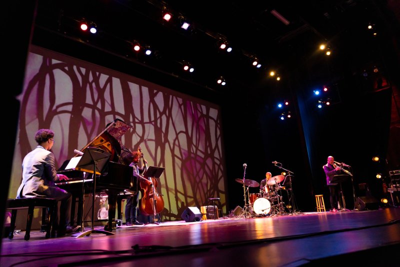 A jazz band performs on a dark stage with a large screen displaying abstract white and purple tree-like patterns behind them.