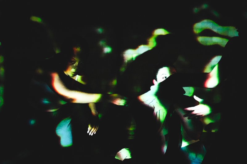 Two dancers in a dark space, illuminated by green and orange light projections, their bodies in motion.