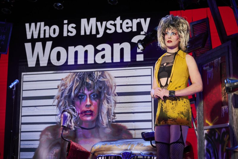 Drag performer in a yellow dress and blonde wig stands before a "Who is Mystery Woman?" poster featuring a mugshot of the same performer.