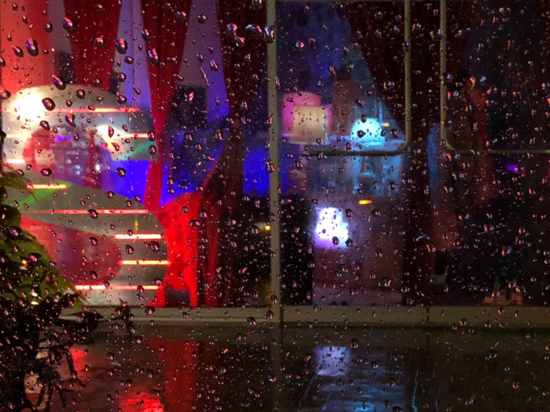 Rain-streaked window with blurred reflections of vibrant red, blue, and purple lights from inside a building.