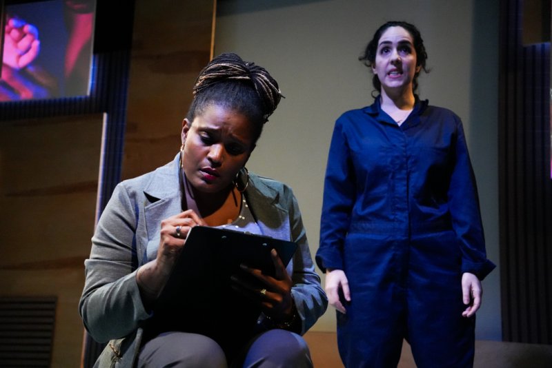 Two women on stage. One sits, writing on a clipboard, wearing a gray blazer. The other stands, looking up, in a blue jumpsuit.