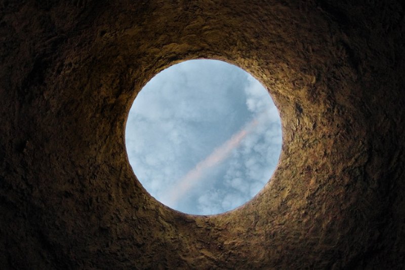 View from inside a dark, textured hole looking up at a bright blue sky with white clouds and a faint pink streak.