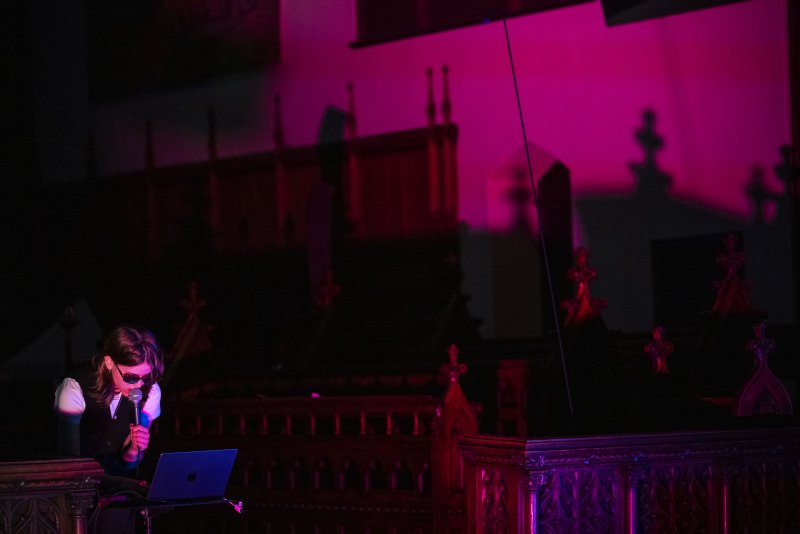 Performer in sunglasses and vest sings into a microphone, illuminated by a laptop and vibrant pink light in a dark, ornate church.