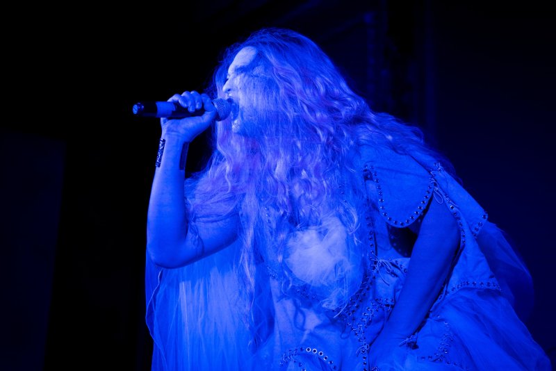 A woman with long, wavy hair sings into a microphone, illuminated by a dramatic blue light against a dark background.