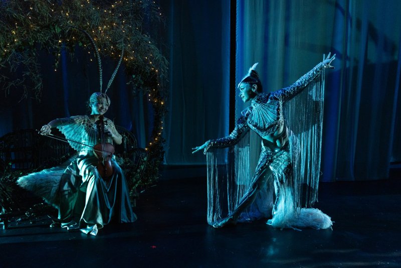 Two performers on stage: one in a dragonfly costume playing a violin, the other in a shimmering outfit dancing, against a dark blue backdrop.