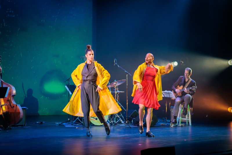 Two dancers in yellow coats, one in black, one in red, perform on a dark stage with musicians and a green-blue backdrop.