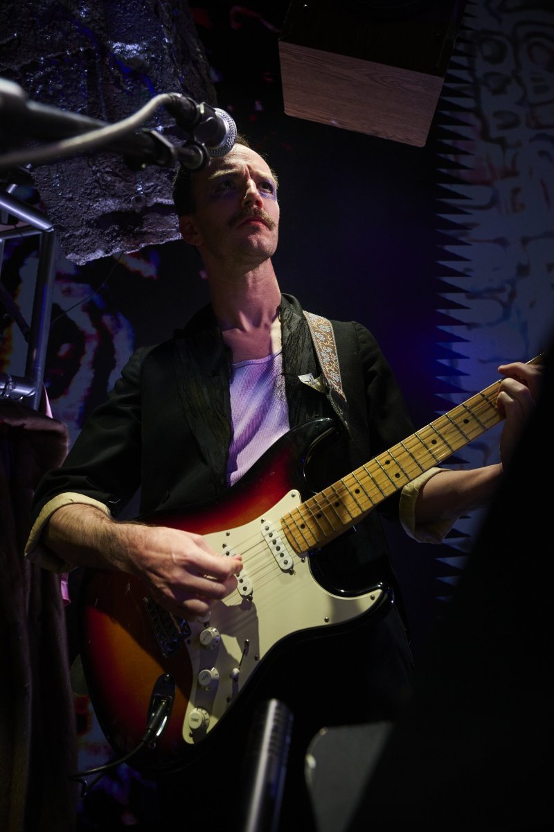 A male musician with a mustache and purple eyeshadow plays a red and white electric guitar under dark lighting, looking upwards.