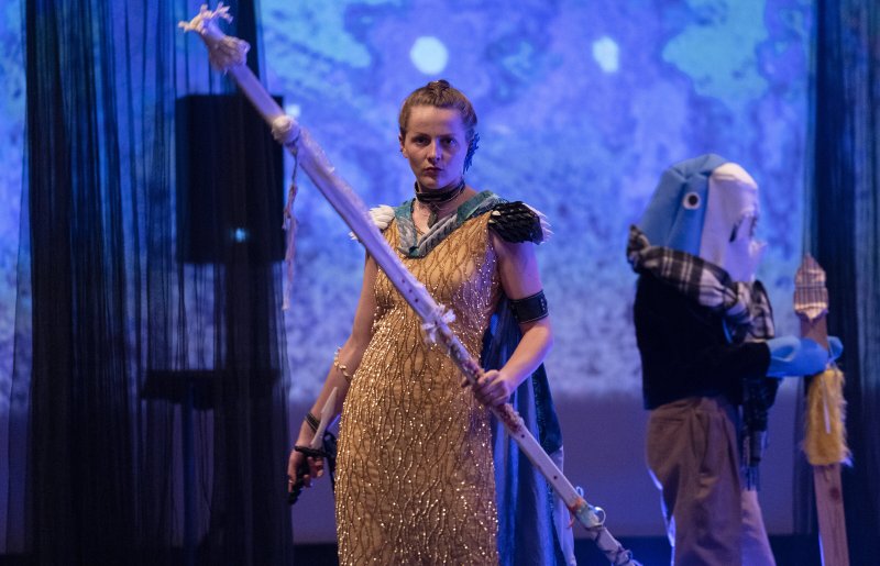 Woman in gold dress and dark cape holding a staff, with a person in a blue shark costume in the background.