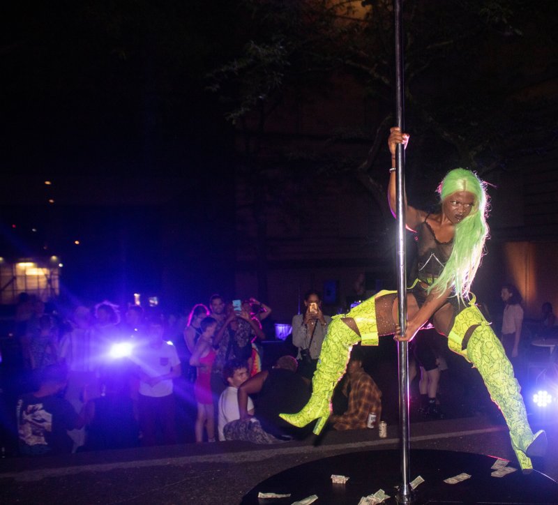 Night scene of a performer with green hair and snakeskin boots on a pole, surrounded by a crowd. Stage is littered with money. Blue and purple lights illuminate the onlookers.