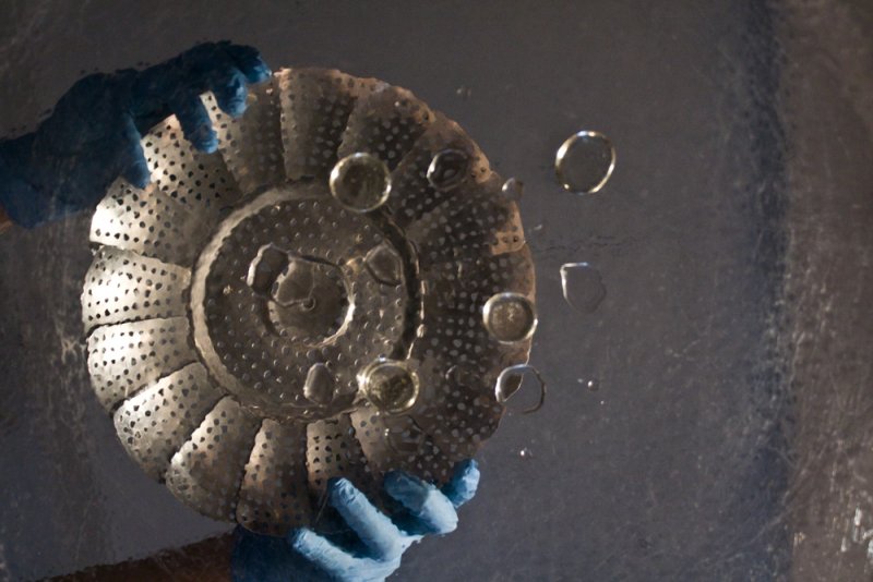 Two gloved hands hold a metal steamer basket underwater, with air bubbles rising against a dark, textured background.