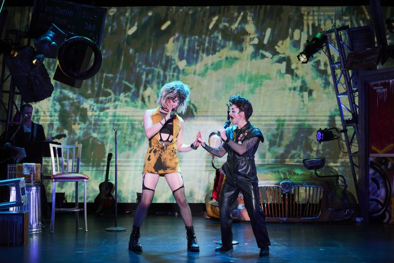 Two performers, one in a yellow dress and the other in black leather, sing into microphones on a stage with a grunge backdrop.