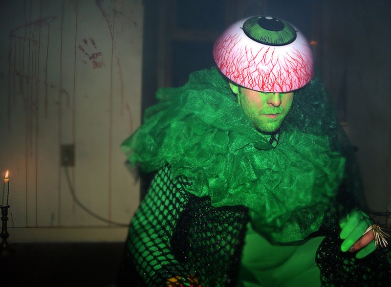 Man in green costume with fishnet sleeves and large eyeball helmet, a lit candle to his left, bloodstains on the wall.