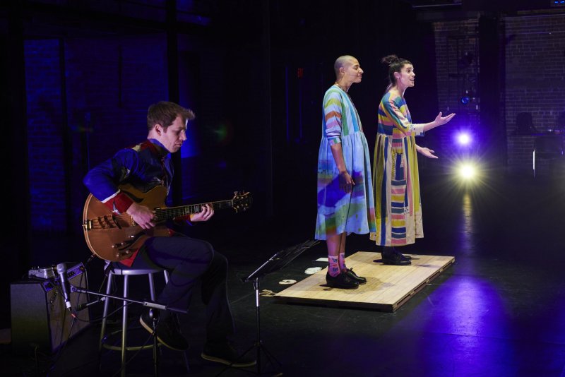 On a stage, a guitarist plays while two singers in colorful dresses stand on a wooden platform, bathed in stage lighting. The backdrop is a dark brick wall.