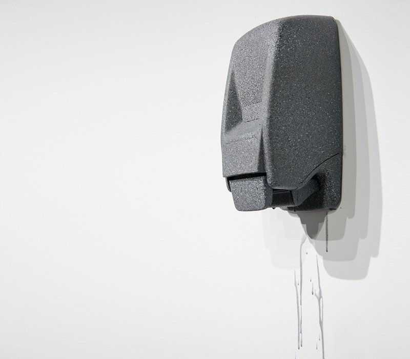 Dark gray, speckled soap dispenser mounted on a white wall, with gray liquid dripping down from its base.