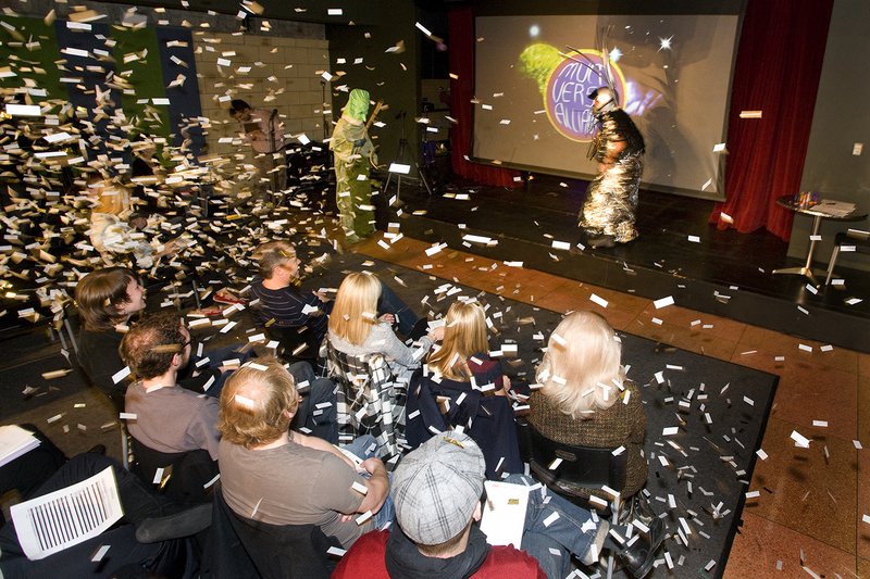Confetti rains on an audience watching two costumed performers on stage with a screen displaying "Multiversal Alliance.