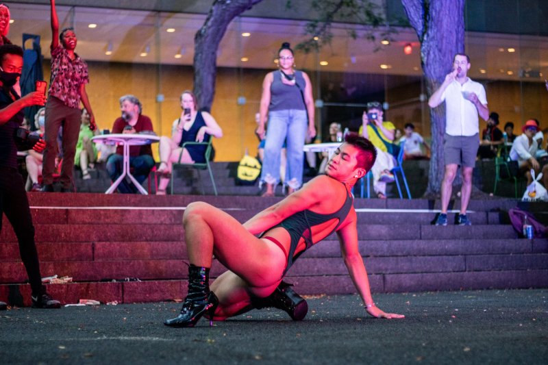 Performance art scene: dancer in black bodysuit and heeled boots poses on ground; audience sits on steps in background. Red and purple lighting.