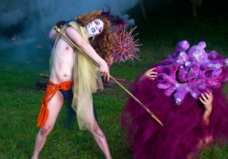 Performer in white face paint and orange sash holding a staff, facing a person in a purple ruffled costume.