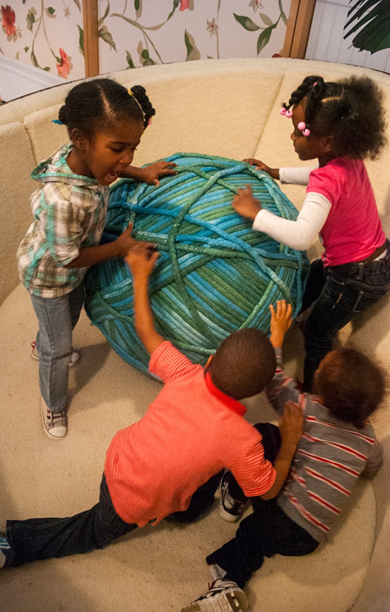 Four young children, two girls and two boys, playing with a large, teal and blue yarn ball on a cream-colored couch.