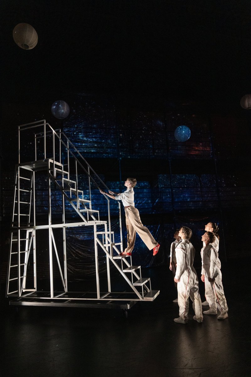 A person in a white shirt and tan pants climbs a white metal staircase, while three people in white jumpsuits watch from below.