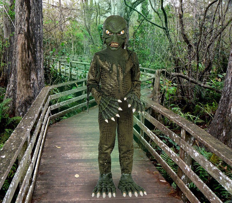 A person in a Creature from the Black Lagoon costume stands on a wooden boardwalk in a swamp.