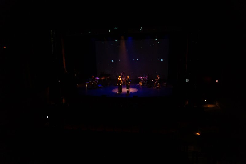 Dark stage with two singers in spotlights, band members, and a starry blue projection on a screen behind them.