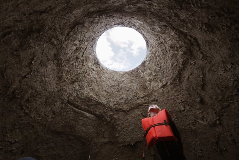 A person in an orange life vest looks up at a circular opening to a cloudy sky from inside a dark, textured well.