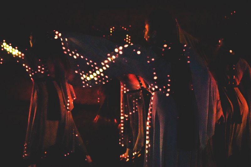 Dark, blurry photo of three dancers in flowing dresses, illuminated by rows of sparkling orange and yellow lights.