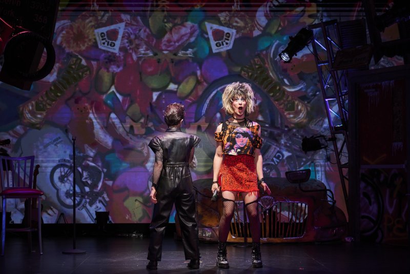 Two performers on stage, one in black leather facing away, the other in a red skirt and blonde wig singing into a microphone.