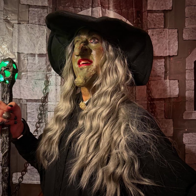 A person dressed as a witch with green skin, red lips, a black hat, and long gray hair, holding a glowing green staff.