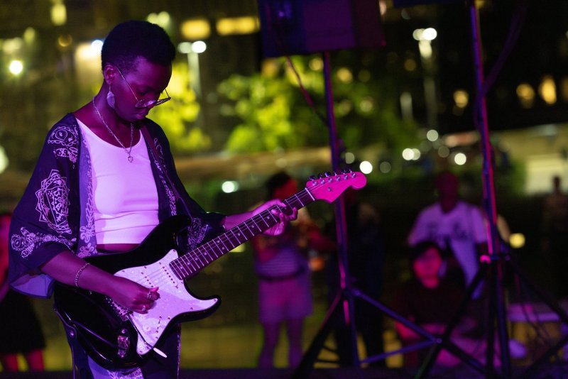 A musician plays an electric guitar under purple stage lights. She wears a patterned kimono, white top, and glasses. Background shows blurred figures and city lights.