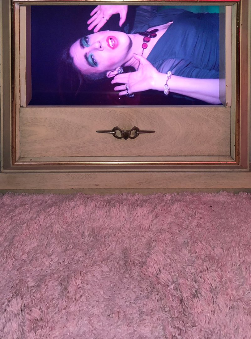 A person with dramatic makeup and dark clothing, illuminated by purple light, displayed on a screen set into a wooden drawer.