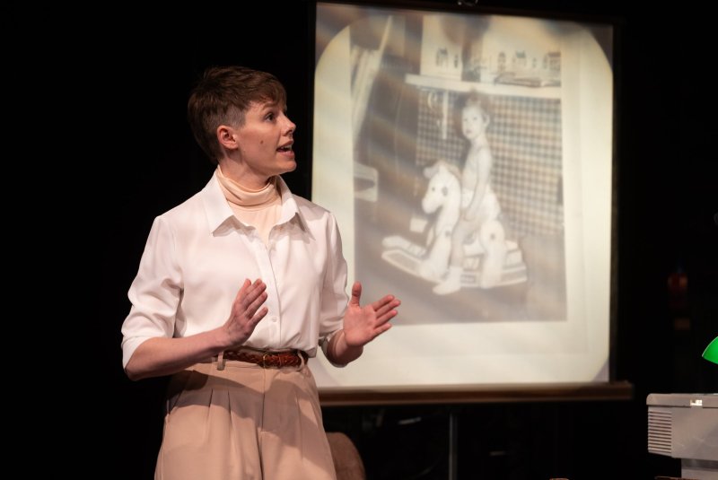 Woman in white shirt and tan pants gestures while speaking; behind her, a projected black and white photo of a child on a rocking horse.