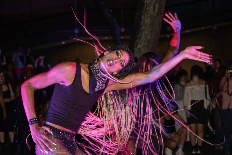 Two performers in drag, lit with pink and blue, strike dynamic poses. Braided hair whips through the air. One wears a black tank top and studded bandana. A crowd is visible in the background.