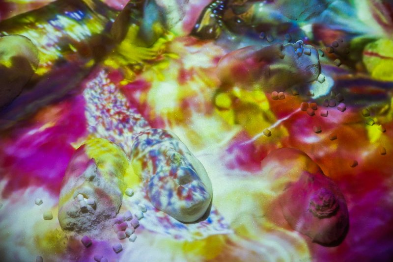 Close-up of a textured surface with small rocks and cubes, illuminated by a vibrant projection of pink, yellow, and white abstract patterns.