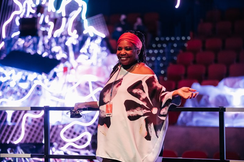 Smiling woman in a red headwrap and floral top, standing before glowing white neon art in a dark room with red seats.