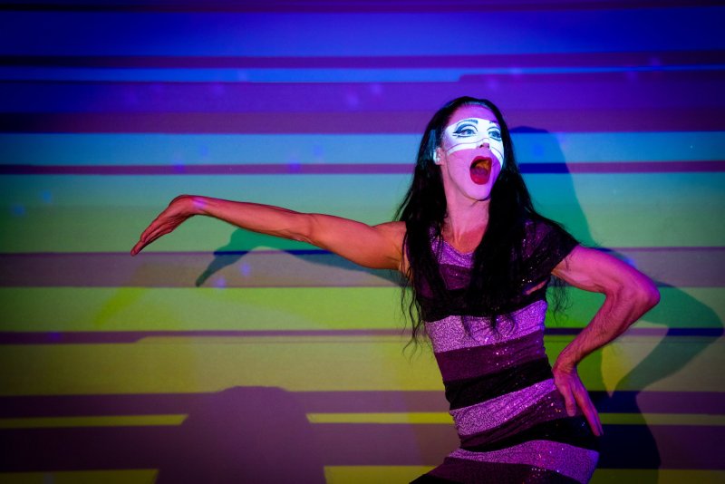 Performer in drag with white face paint and open mouth, wearing a striped sequin dress, against a background of colorful horizontal stripes.