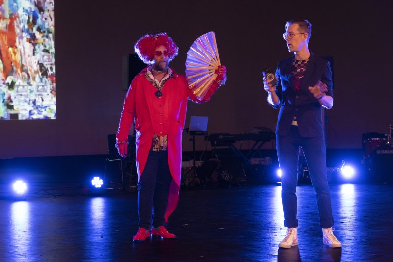 Two performers on stage: one in a red wig, coat, and shoes, holding a fan; another in a suit and glasses.