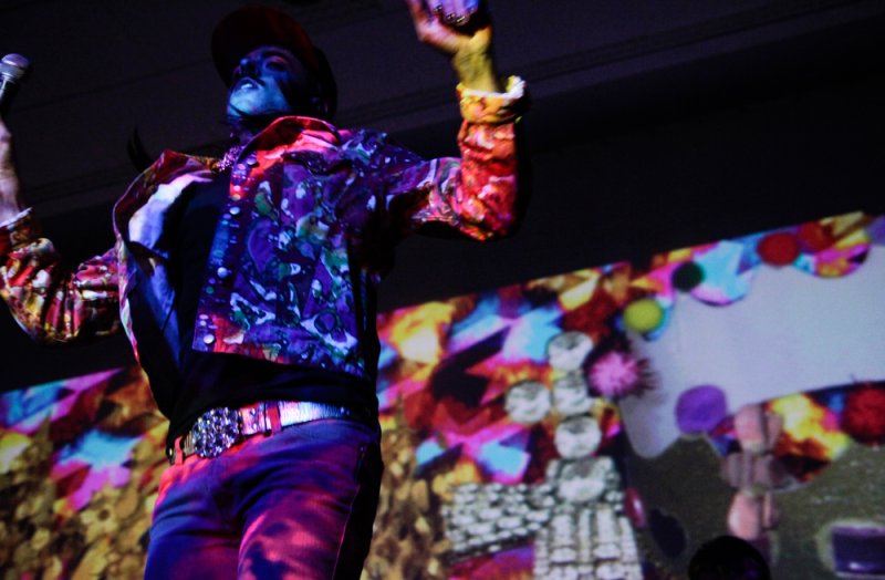 Performer in a colorful jacket and hat, with blue-painted face, on stage with vibrant projected visuals.
