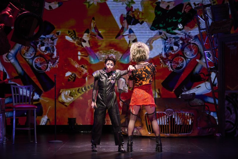 Two performers on stage, one in black leather and the other in a ripped orange dress, against a vibrant red backdrop with abstract art.