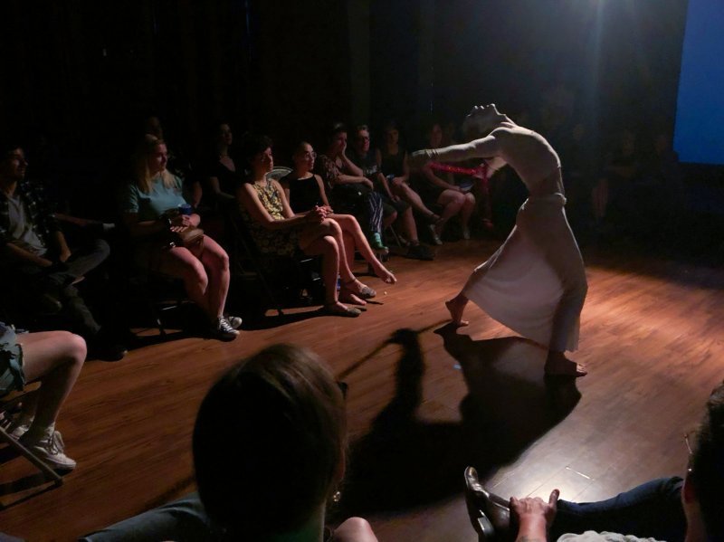 Performance art piece: a figure in white bends backwards on a wood stage, holding a red object. An audience sits in the dark, watching. Spotlight on the dancer.